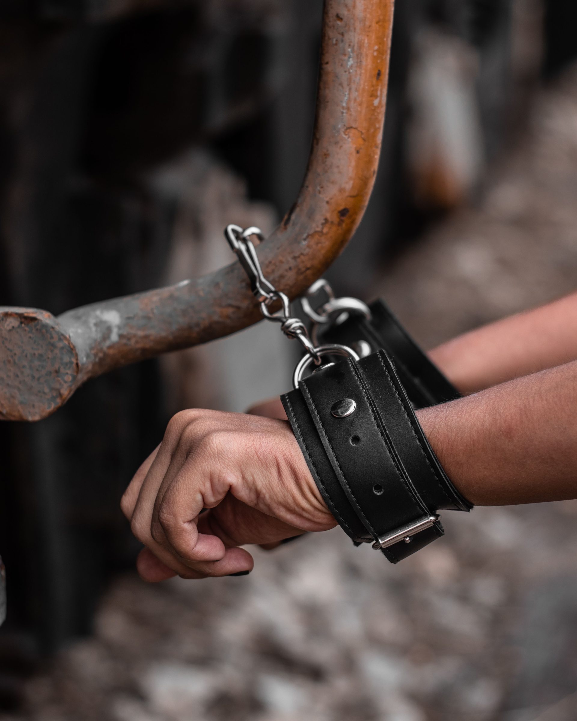 A detailed view of hands in black leather handcuffs attached to a rusty rail outdoors.